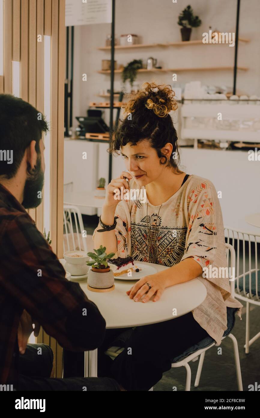 Gioioso casual Donna mangiare torta con forchetta mentre si siede al tavolo in caffetteria guardando il suo ragazzo Foto Stock