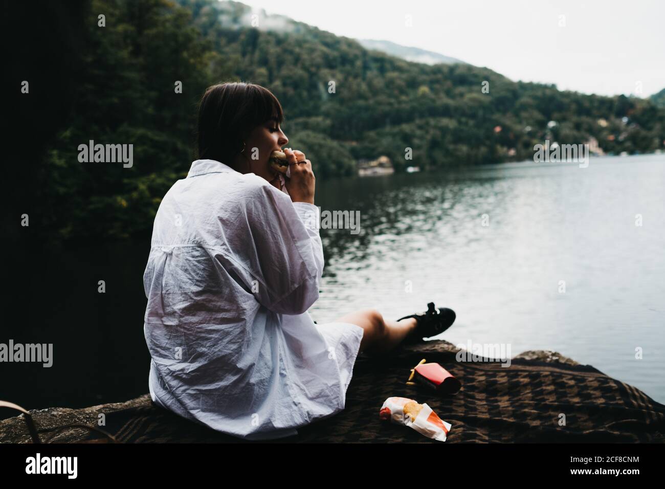 Donna che mangia hamburger vicino al lago e alle montagne Foto Stock