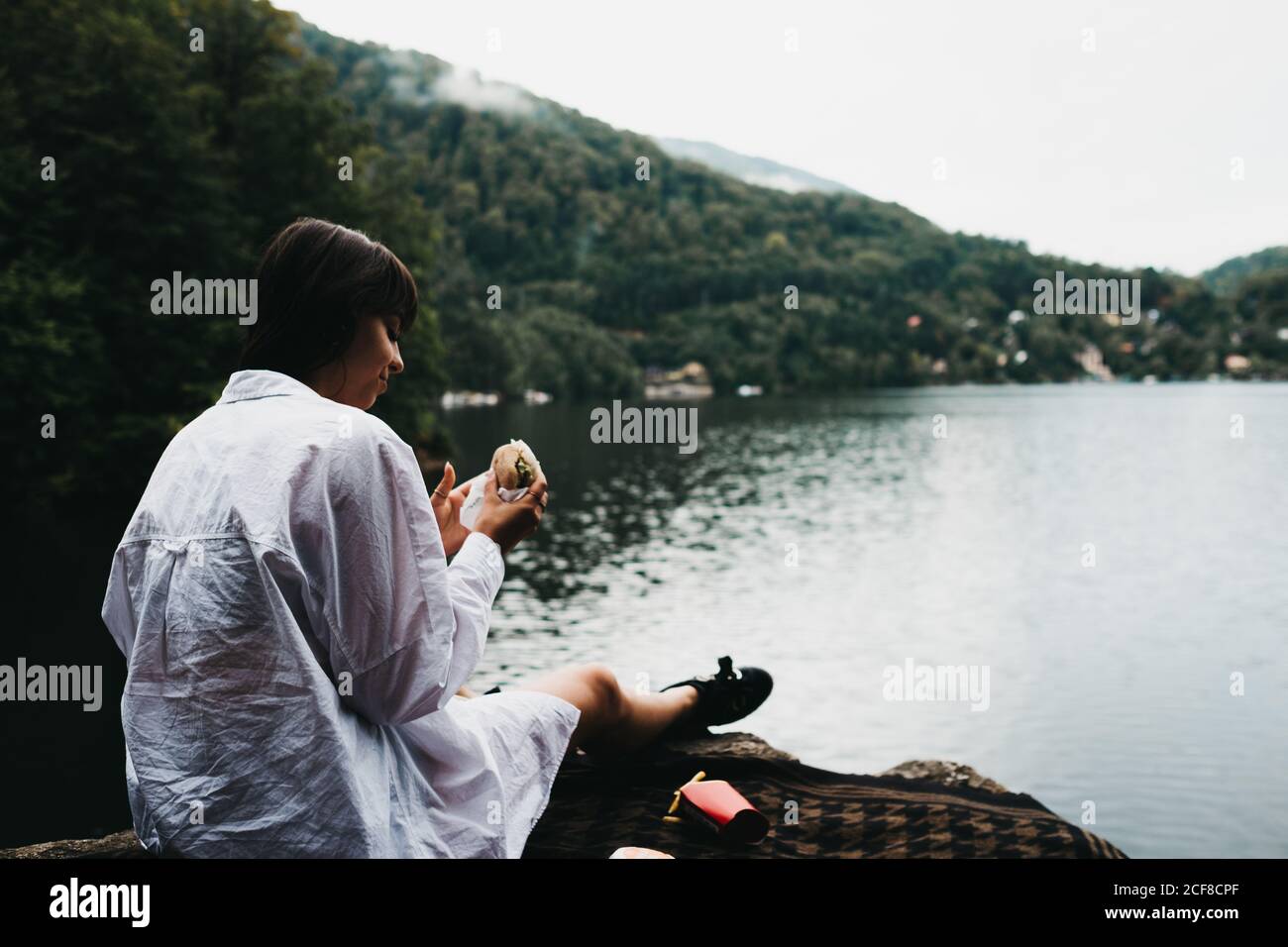 Donna che mangia hamburger vicino al lago e alle montagne Foto Stock