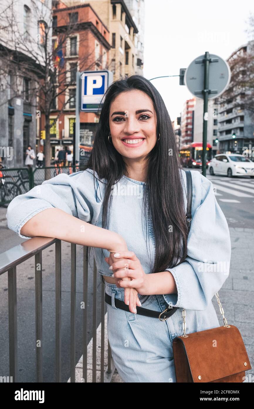Giovane donna bruna ottimista in outfit elegante con borsetta appoggiata gomito su recinzione di metallo mentre si sta in piedi sulla strada della città e. guardando la fotocamera Foto Stock