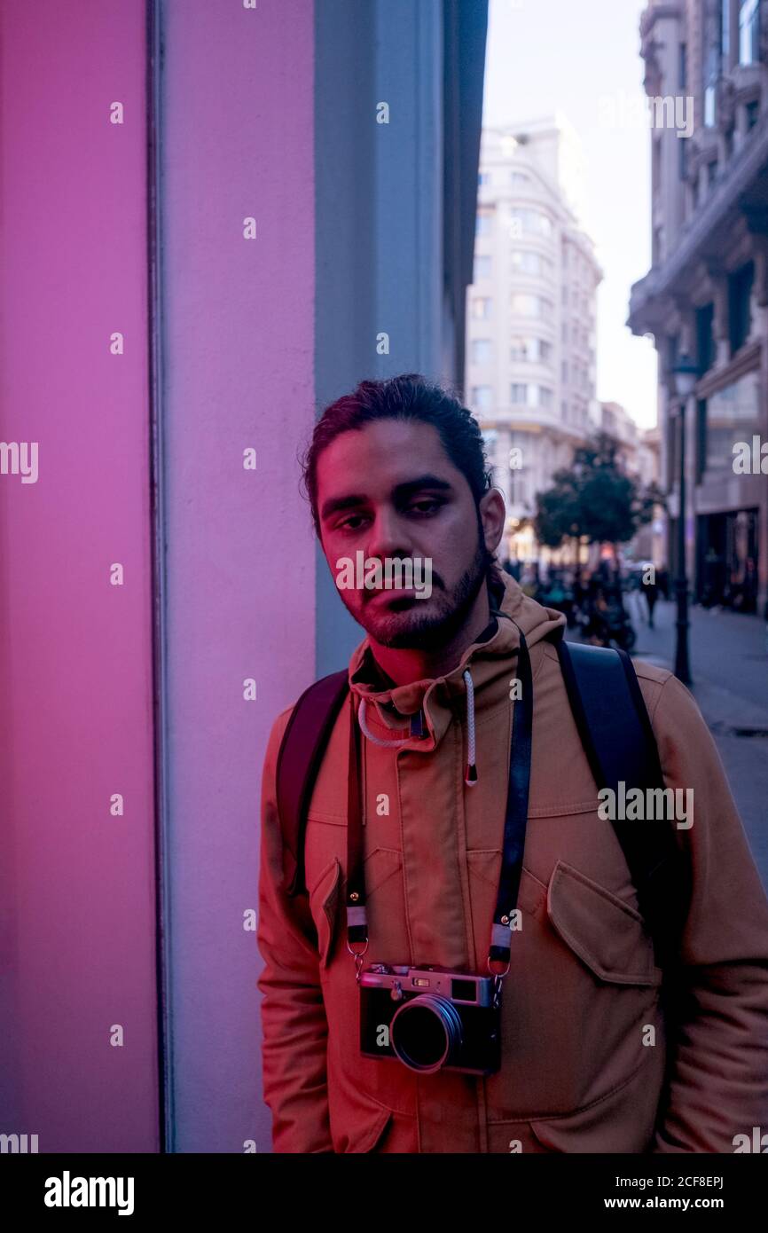 Bel fotografo etnico maschile in abiti casual con zaino nero e un panino di capelli in piedi sulla strada vicino alla vetrina con bagliore al neon e videocamera con videocamera per riprese appeso al collo Foto Stock