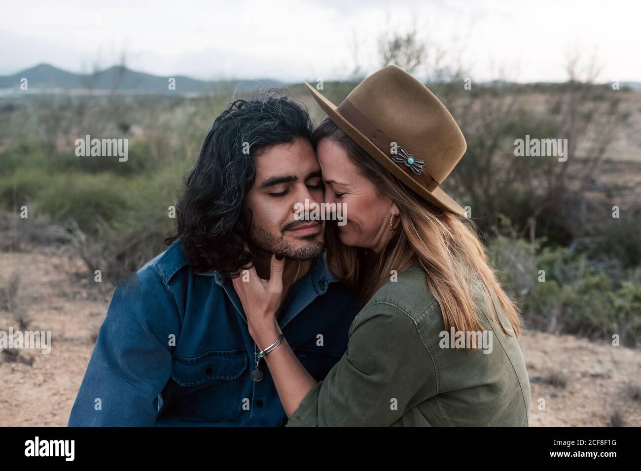 uomo in cappello seduto su terreno sabbioso e abbracciando felice ragazza durante la data romantica in serata nuvolosa in campagna Foto Stock