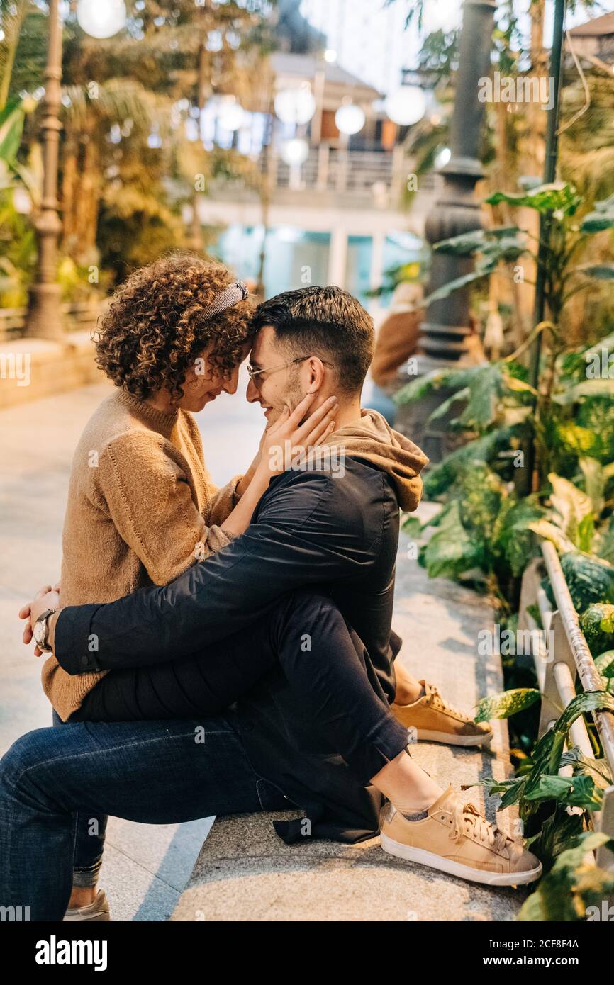 Vista laterale del giovane uomo e della donna che abbraccia e tocca la fronte mentre si siede sulla panchina durante la data romantica sulla strada della città Foto Stock