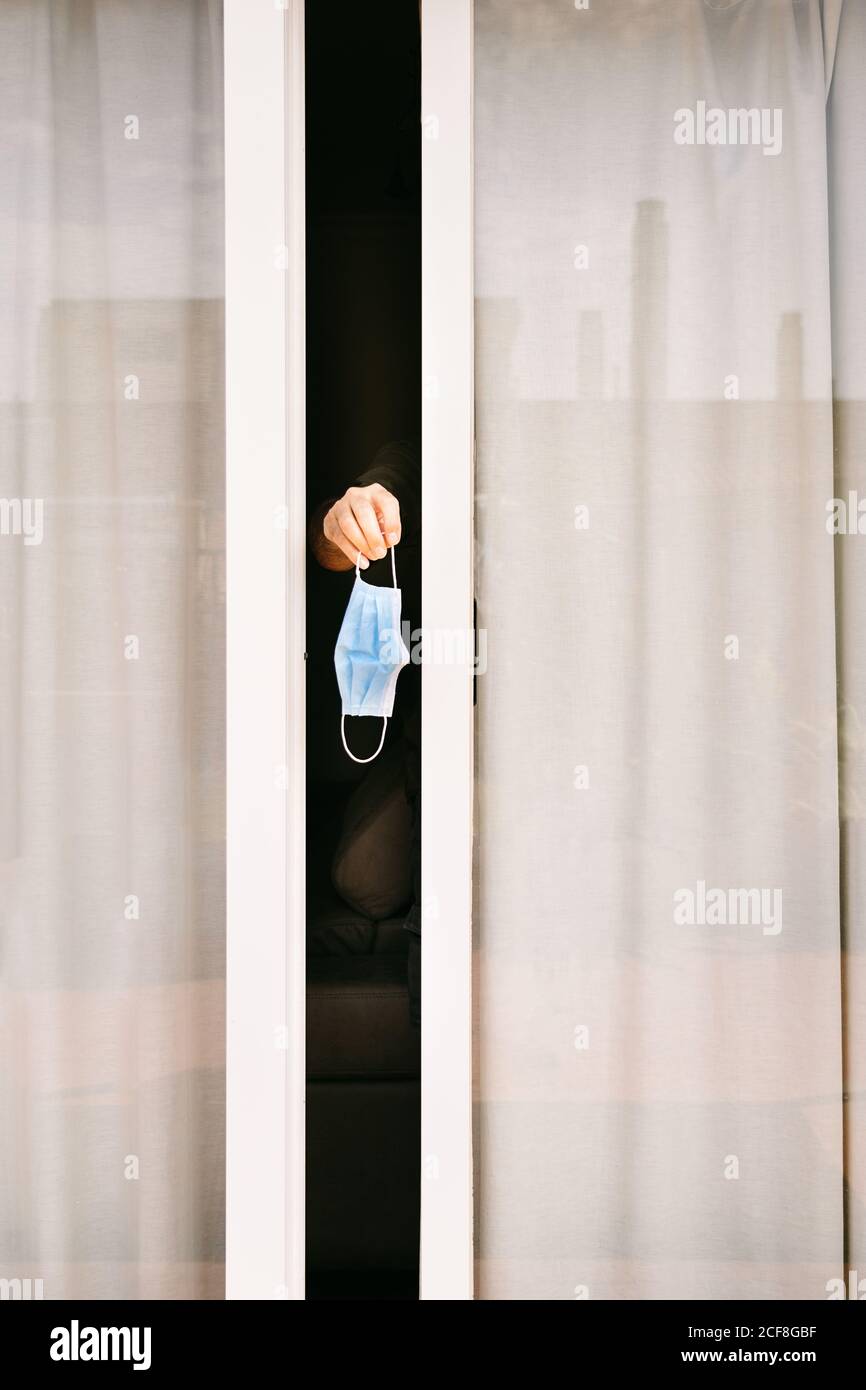 Mano di persona anonima che esce dalla finestra leggermente aperta tenuta della maschera protettiva che rimane in quarantena durante l'epidemia di coronavirus Foto Stock