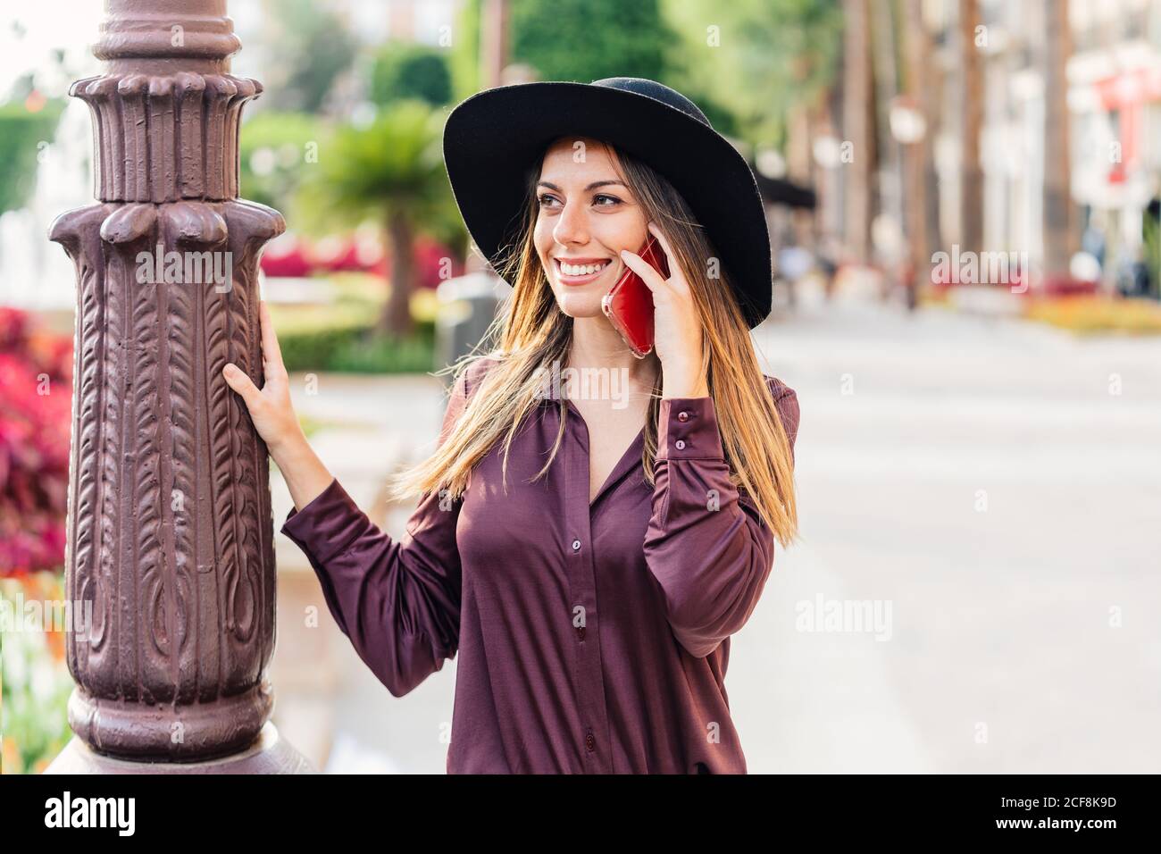 Contenuto long haired lady in cappello nero alla moda e camicia camminando sulla strada mentre si chiama sul telefono cellulare e. vista sulla strada della città Foto Stock