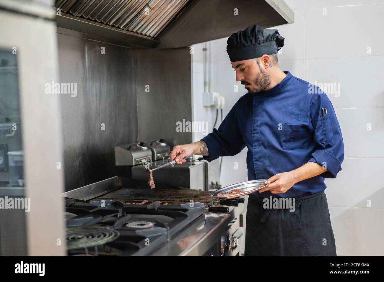 Cuoco maschile in grembiule nero e cappello che tiene in mano piastra metallica e pinze e fette di carne alla griglia piano cottura mentre si lavora nella cucina del ristorante Foto Stock