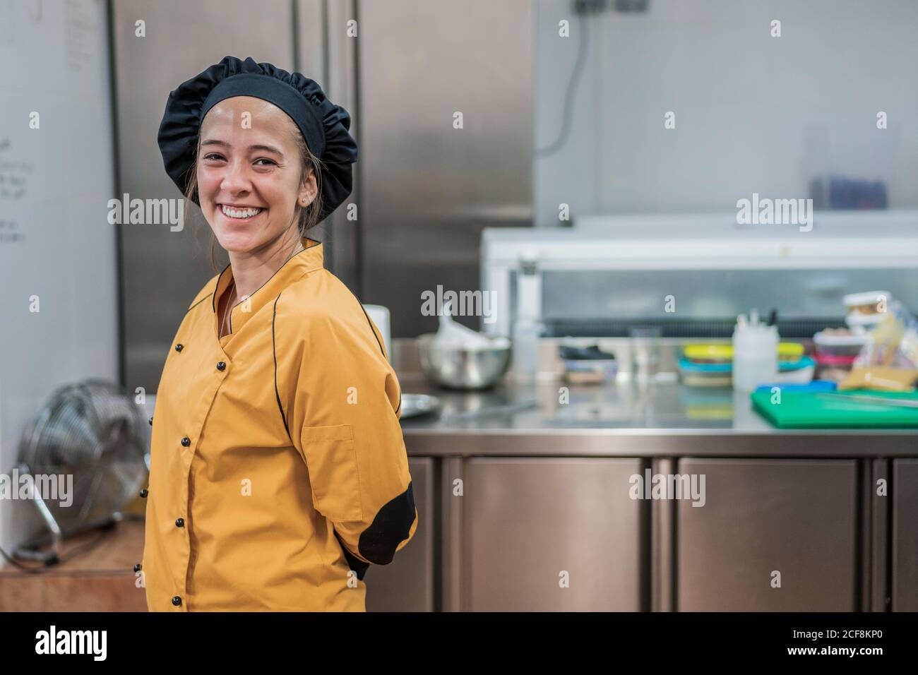 Giovane cuoca allegra in uniforme gialla e cappello nero guardando la macchina fotografica mentre si è in piedi nella cucina del ristorante Foto Stock