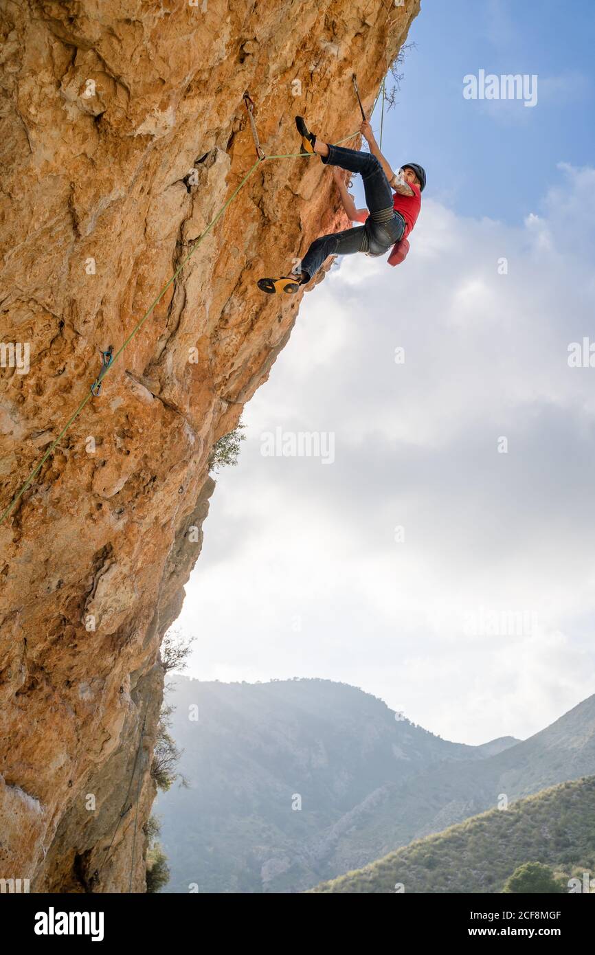 Dal basso alpinista sportivo femminile ascendente sulla scogliera in montagna terreno in giorno nuvoloso Foto Stock