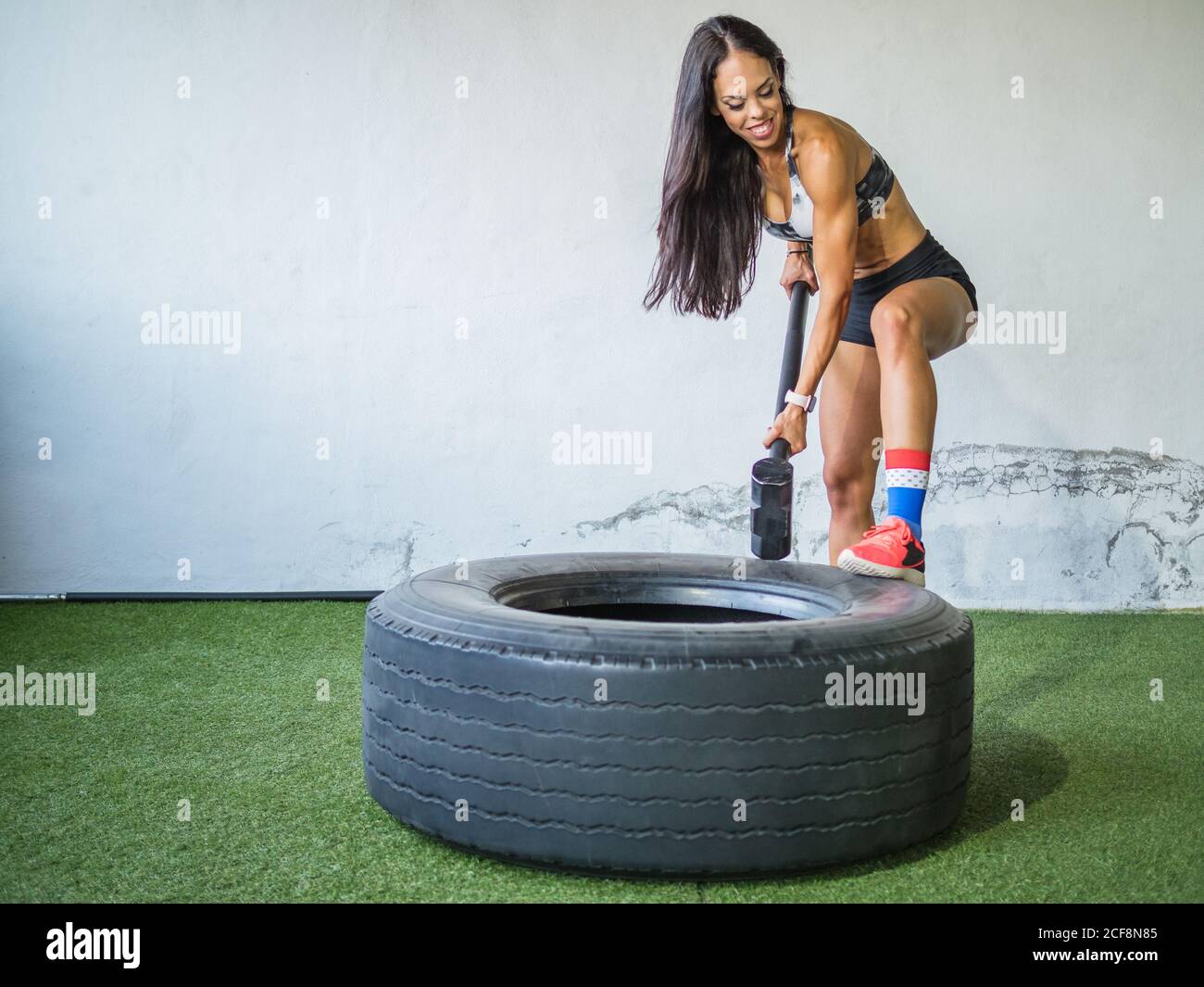 Sicuro allegro forte atleta adulta in sportswear mettendo a fuoco mentre lavorare con un martello pesante e uno pneumatico su un tappeto verde contro la parete bianca nel club sportivo Foto Stock