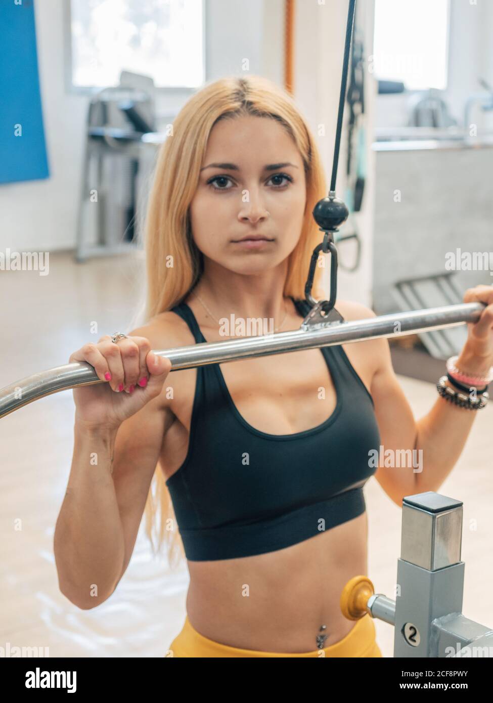 Slim sportswoman che tira giù la barra della macchina di esercitazione durante l'addestramento in palestra che guarda la macchina fotografica Foto Stock