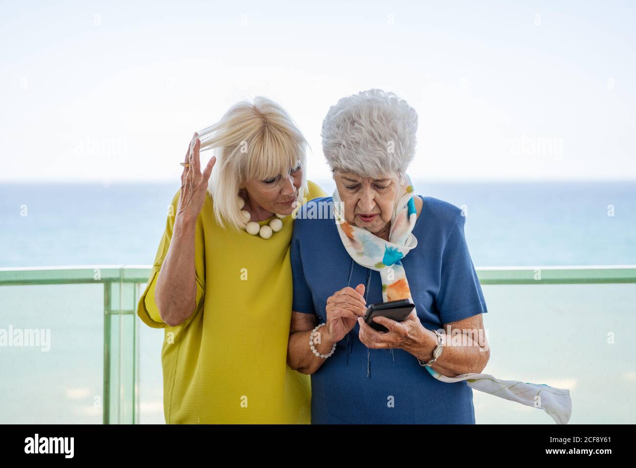 Vecchi amici in abiti eleganti che navigano insieme lo smartphone mentre si levano in piedi e riposandosi sul balcone contro il mare sul resort Foto Stock