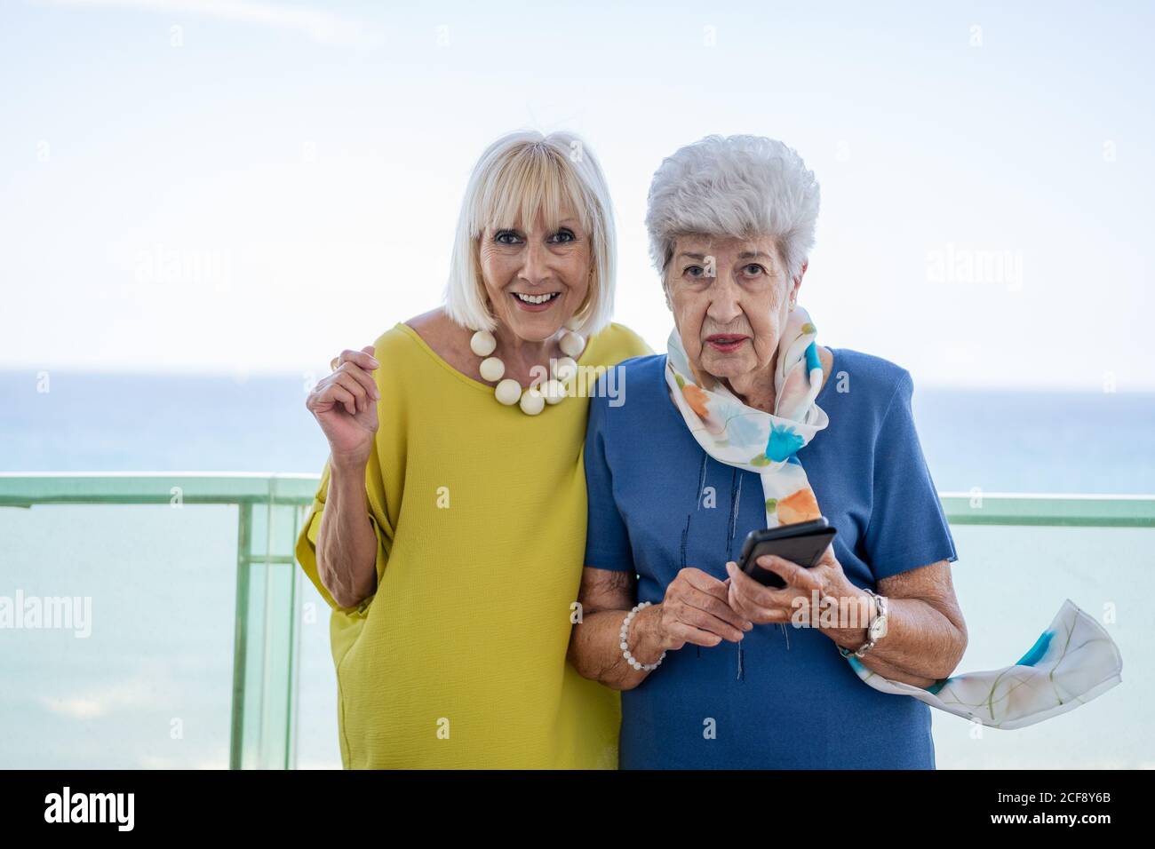 Vecchi amici in abiti eleganti che navigano insieme lo smartphone mentre si levano in piedi e riposandosi sul balcone contro il mare sul resort Foto Stock