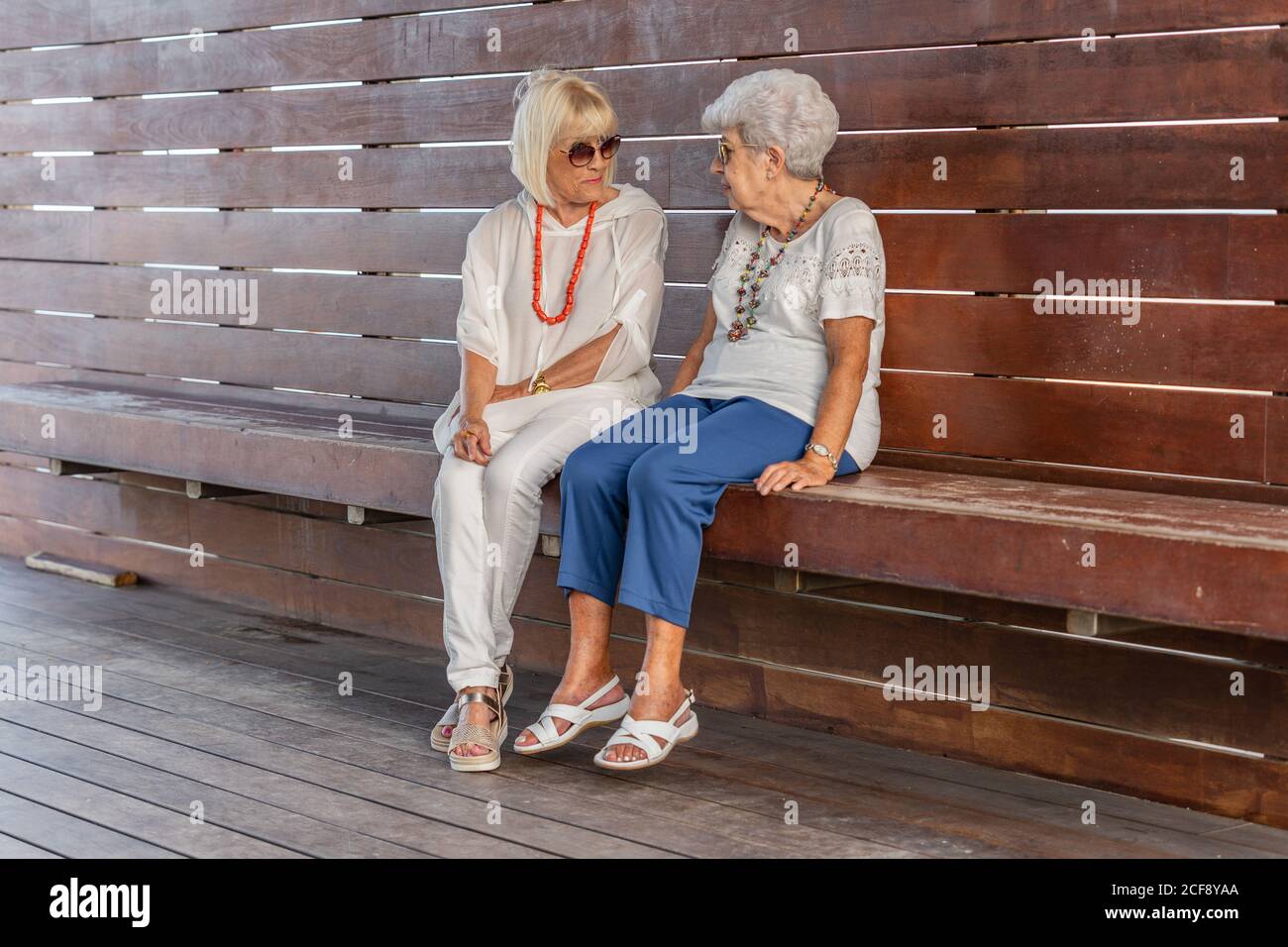 Sicuro alla moda allegro grigio-capelli donne senior in abiti estivi e. occhiali da sole che parlano su una grande panca di legno che si guarda l'un l'altro Foto Stock