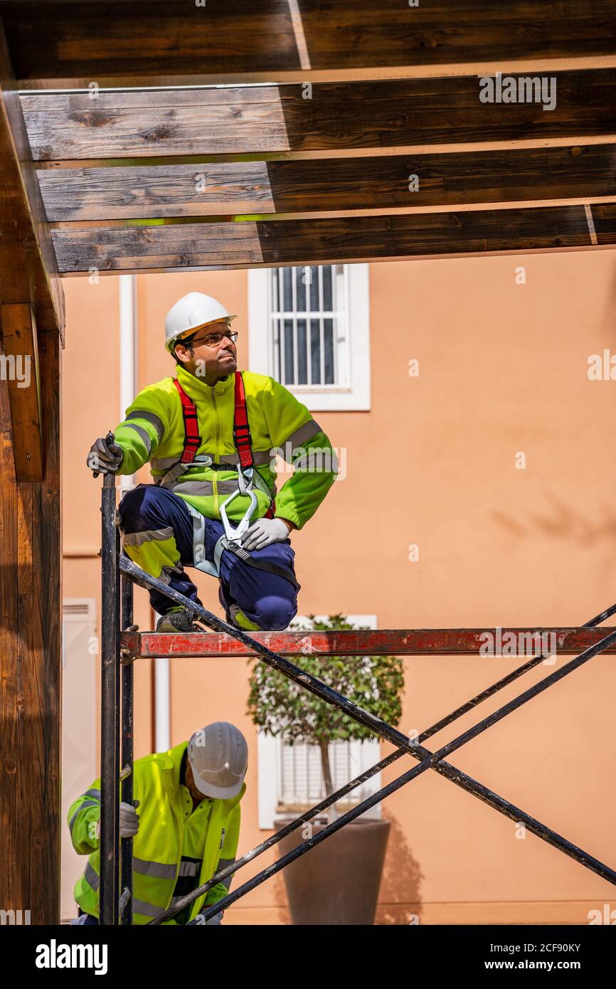 Ingegnere professionista in uniforme e casco seduto su ponteggi e. guardare via mentre si lavora con un collega sulla manutenzione di elettricità attrezzature vicino a un edificio residenziale Foto Stock