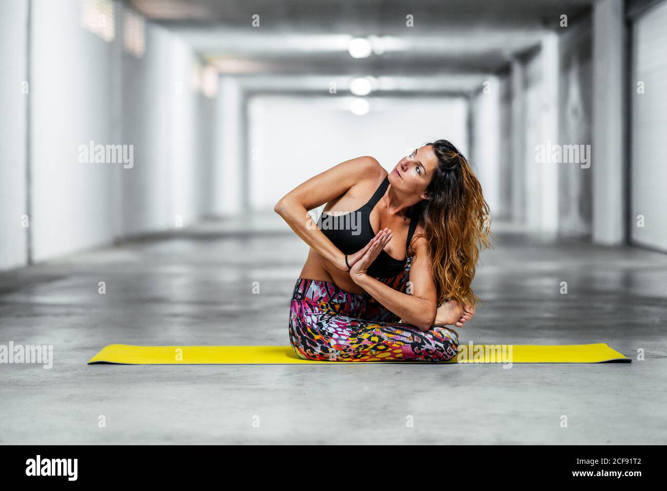 Vista laterale della giovane femmina concentrata in usura attiva seduta twist asana con le mani di preghiera mentre si pratica yoga in garage leggero Foto Stock
