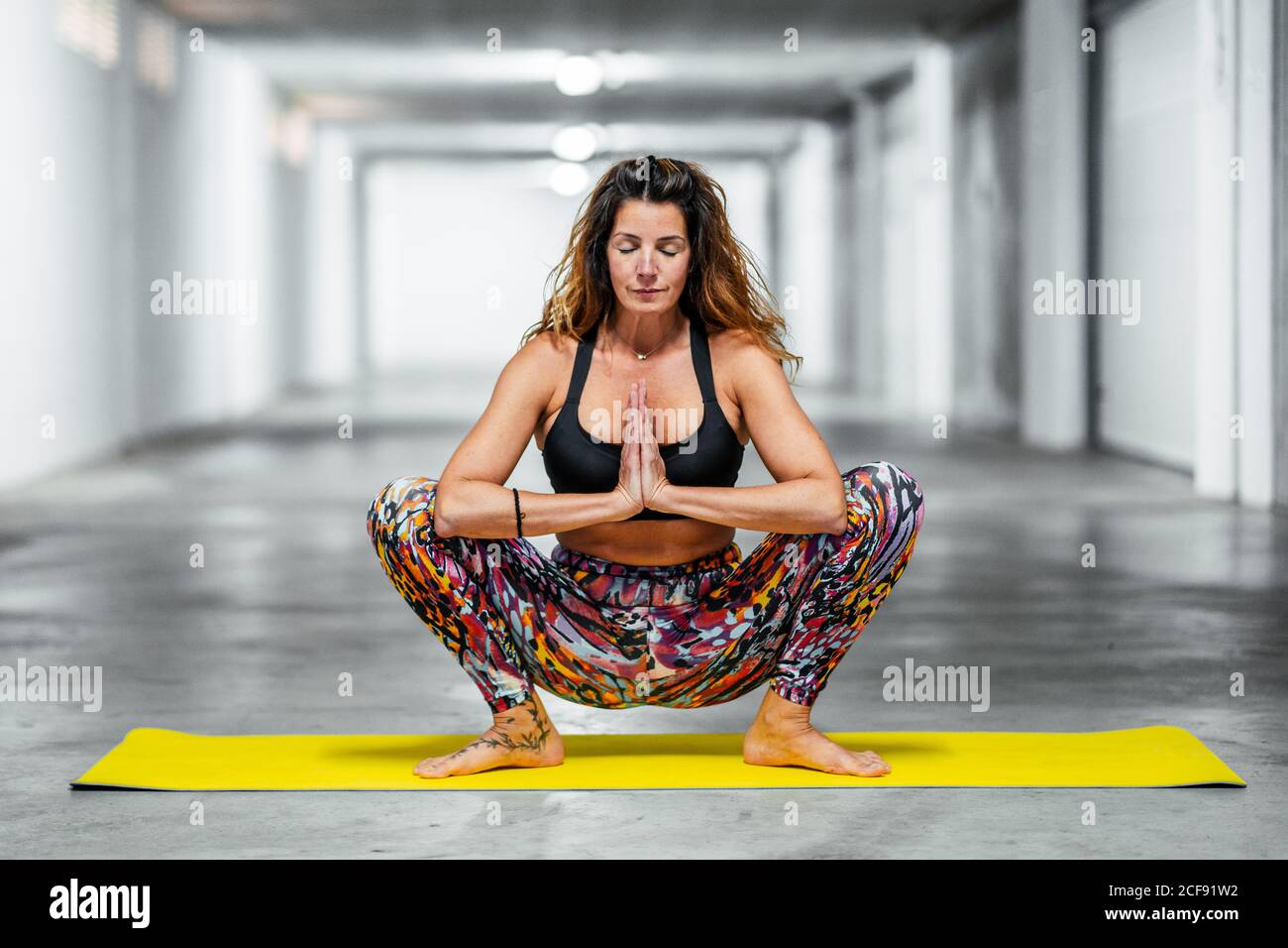 Giovane donna concentrata in reggiseno sportivo e leggings seduta dentro Garland posa o Malasana e meditare durante la pratica di yoga in garage Foto Stock