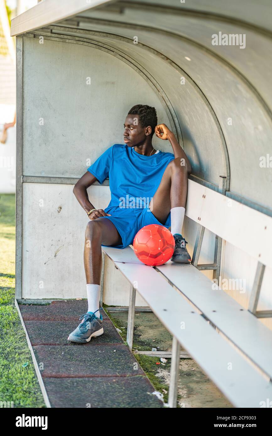 Adolescente afroamericano a corpo pieno con palla di calcio seduta panca e guardare via durante l'allenamento all'aperto Foto Stock