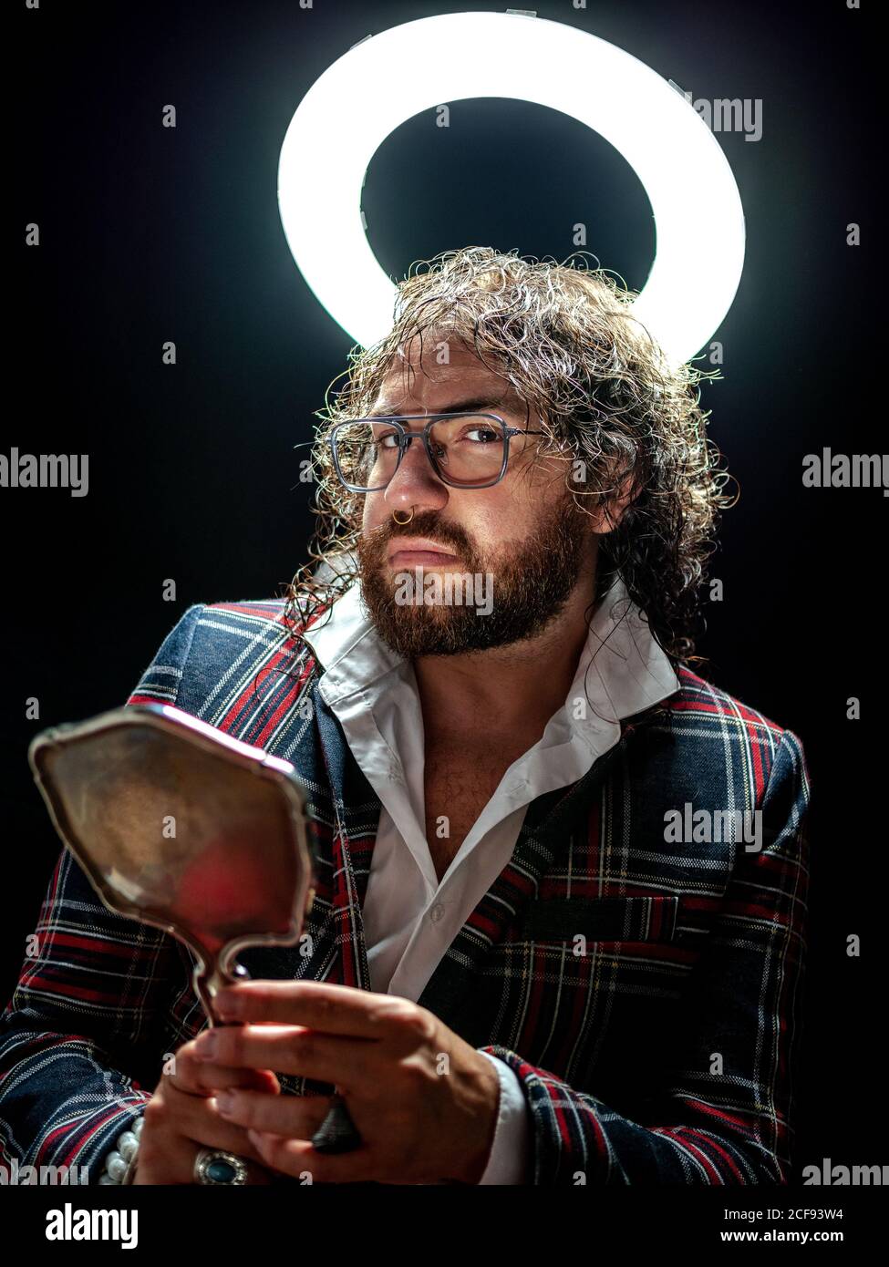 Uomo elegante e ben vestito con la barba che tiene lo specchio di bellezza e. guardando la fotocamera su sfondo nero sotto brillante falso nimbus Foto Stock