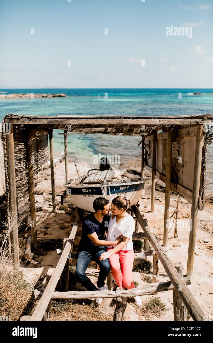 Coppia amorevole coccolarsi vicino barca ormeggiata a terra sotto baldacchino di legno in spiaggia in estate Foto Stock