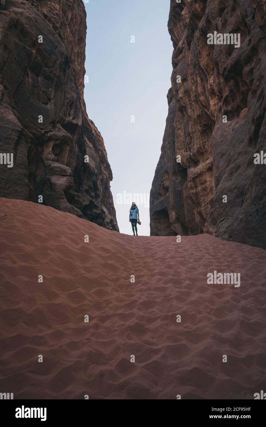 anonimo donna turistica in piedi su sabbia tra pareti di pietra ruvida Contro il cielo blu senza nuvole nella gola del deserto di Wadi Rum In Giordania Foto Stock