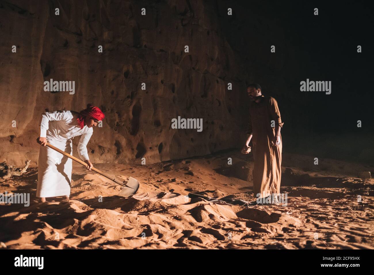 Uomini etnici in abiti arabi tradizionali utilizzando la pala per scavare Sabbia asciutta vicino alla scogliera nel deserto di Wadi Rum Foto Stock