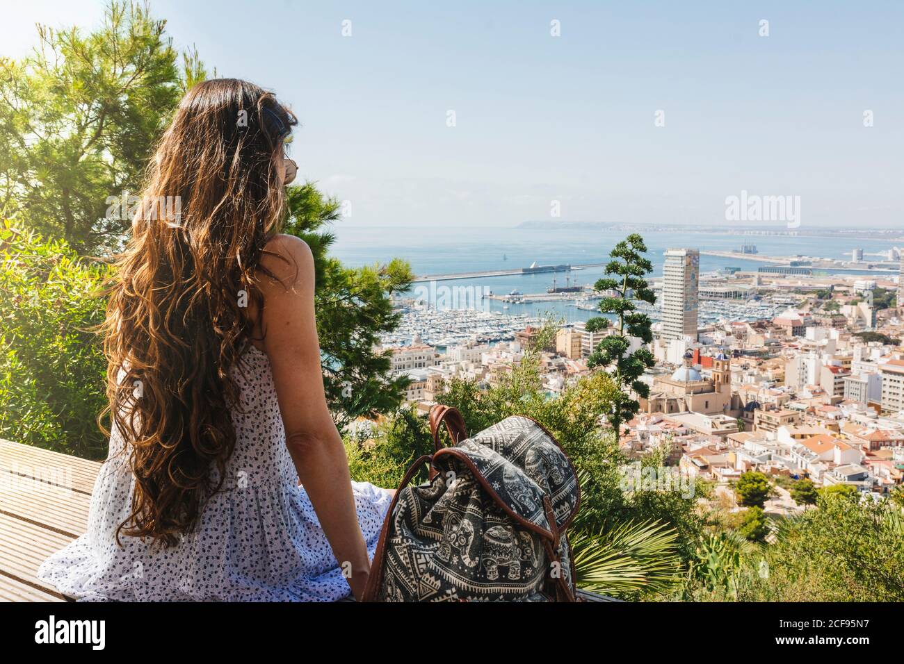 Vista posteriore della giovane donna dai capelli lunghi in vestito estivo con zaino godendo delle viste della città seduto su una panca di legno in giorno luminoso in Alicante Spagna Foto Stock