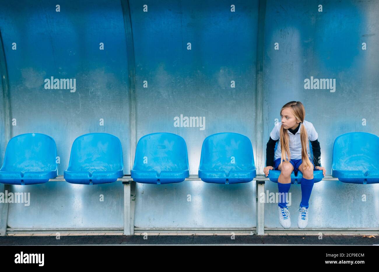 La lunghezza intera ha frustrato la ragazza di preteen in uniforme di calcio che siede da solo sul sedile in plastica blu dopo il fallimento della partita nel club sportivo Foto Stock