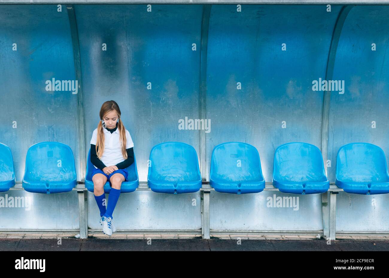 La lunghezza intera ha frustrato la ragazza di preteen in uniforme di calcio che siede da solo sul sedile in plastica blu dopo il fallimento della partita nel club sportivo Foto Stock