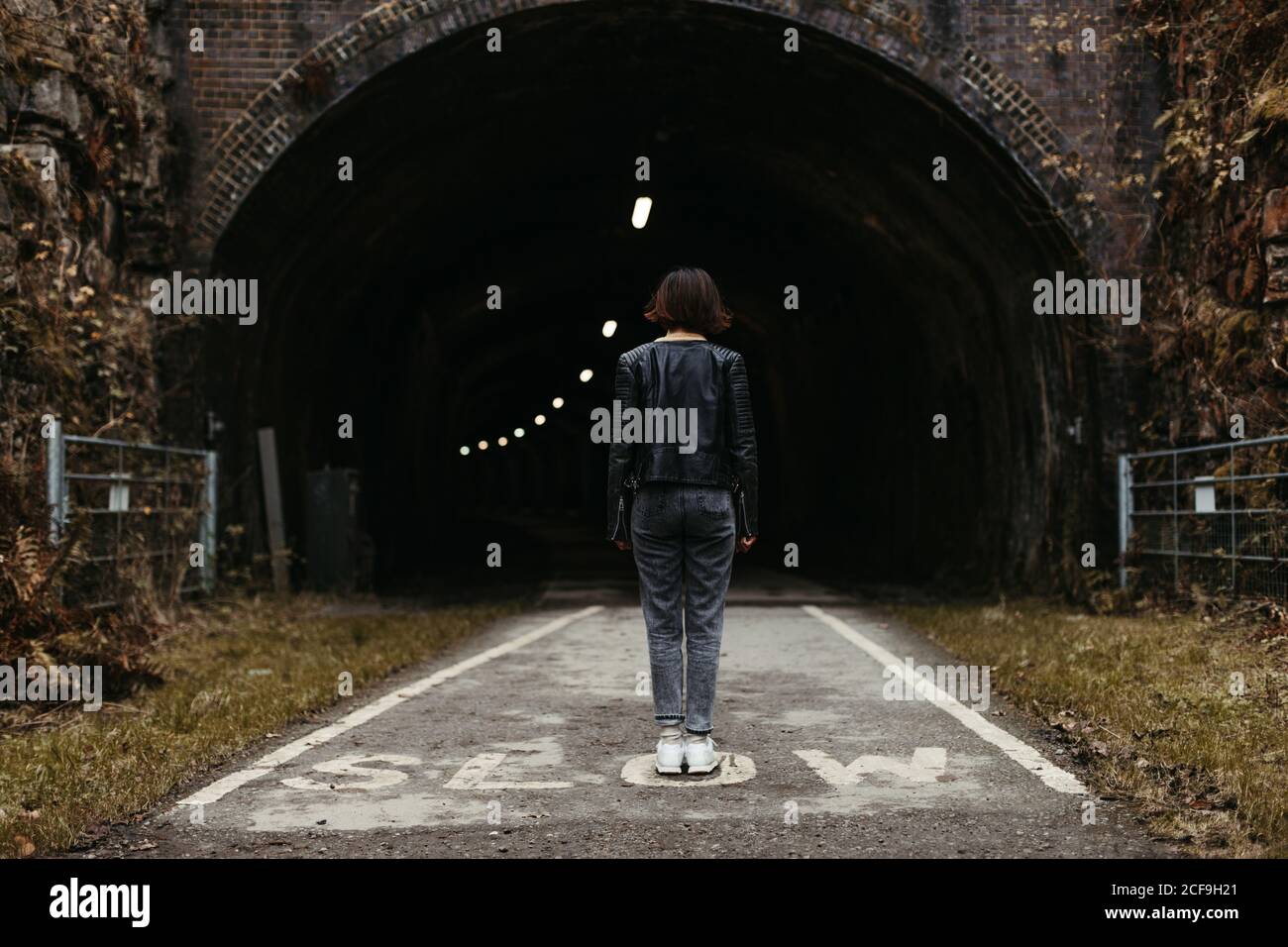Vista posteriore di elegante donna in giacca di pelle nera guardando il tunnel scuro con illuminazione interna su segnato con iscrizione strada lenta nel Regno Unito Foto Stock