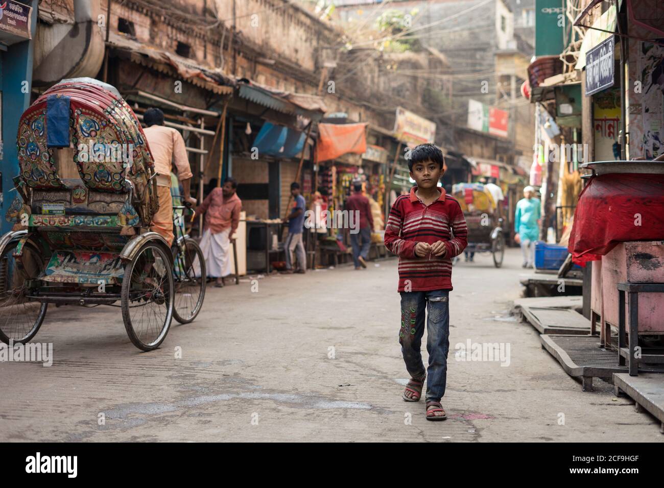 Bangladesh - Gennaio, 25 2019: Povero ragazzo etnico in vestiti sporchi guardando la macchina fotografica mentre cammina su strada asfaltata sul mercato di strada stagionato in città Foto Stock