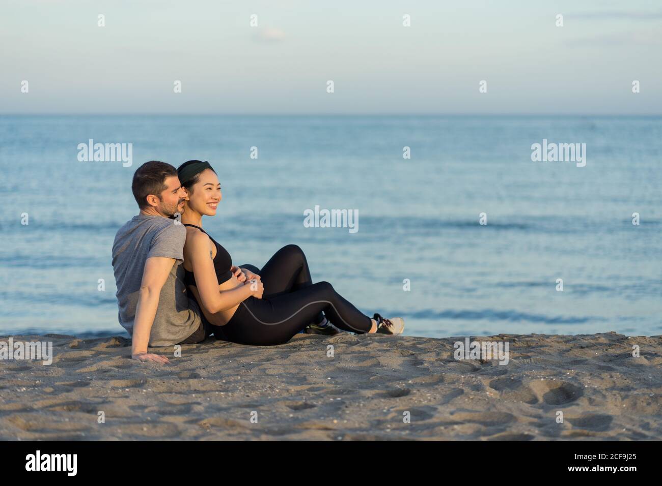 Vista laterale della giovane coppia allegra multirazziale in abbigliamento sportivo seduto sulla spiaggia di sabbia, riposandosi dopo l'allenamento e godendosi del tempo insieme Foto Stock