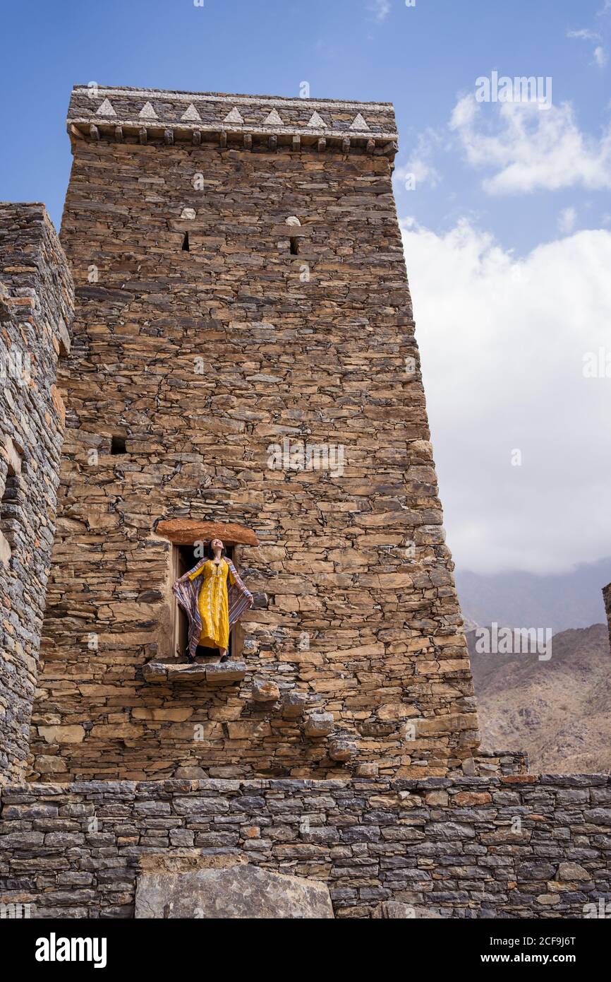 Dal basso di monumentale antico edificio con remota turista femminile uscendo dalla porta in abito giallo mentre godendo caldo Giornata di sole in Marble Village ad al Bahah Foto Stock