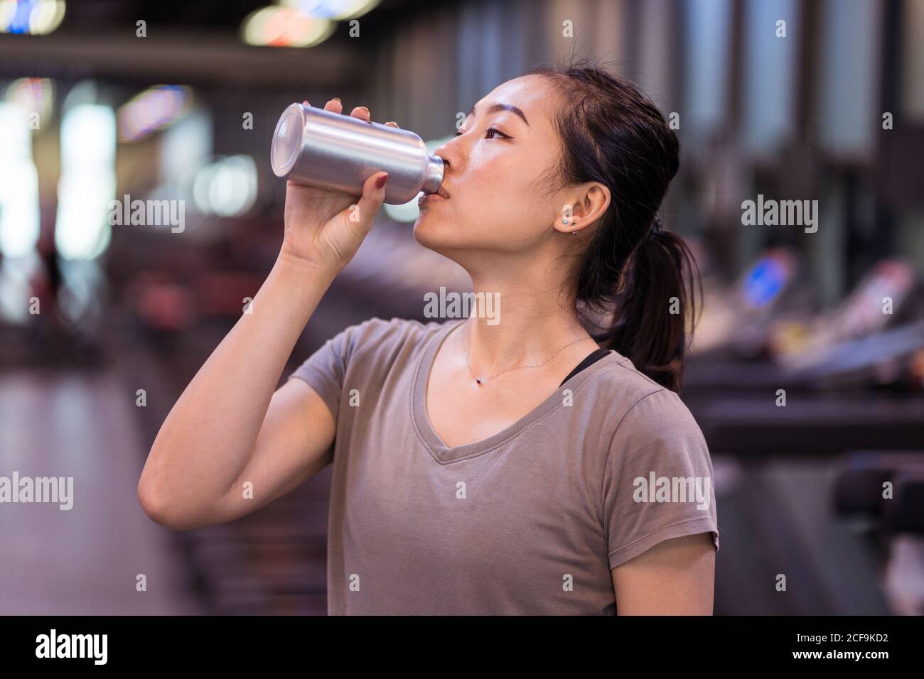 Giovane sportivo asiatico sottile in abbigliamento sportivo bere acqua fresca dalla bottiglia mentre si riposa dopo un duro allenamento in palestra moderna Foto Stock