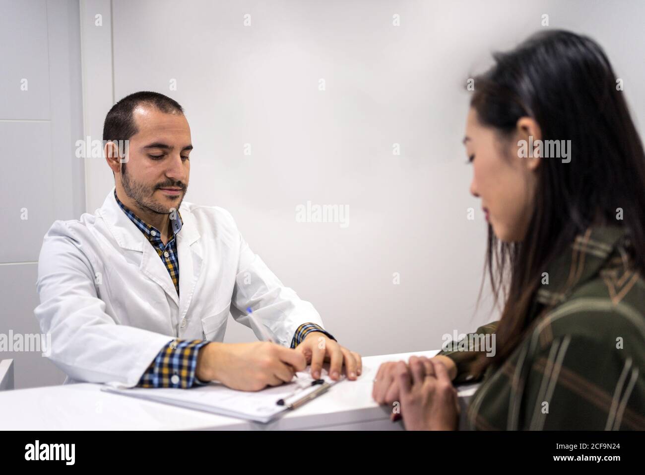 Attento medico maschile che compila il modulo di carta mentre si sta con Paziente femminile asiatica alla reception in ufficio medico Foto Stock