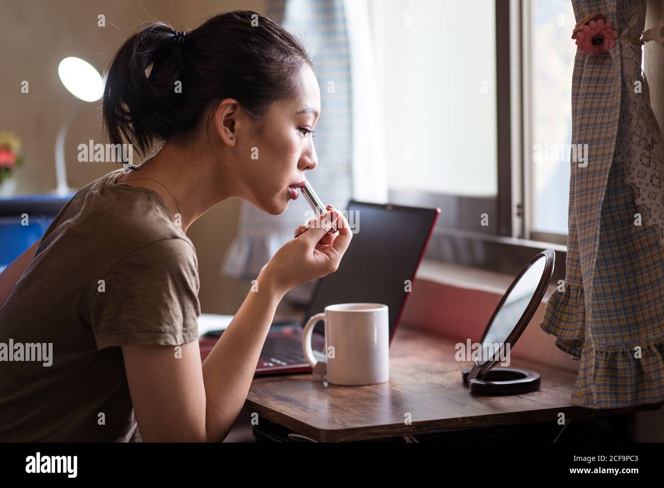 Vista laterale della giovane donna etnica seria in abbigliamento casual sedersi al tavolo davanti allo specchio e applicare il rossetto Foto Stock