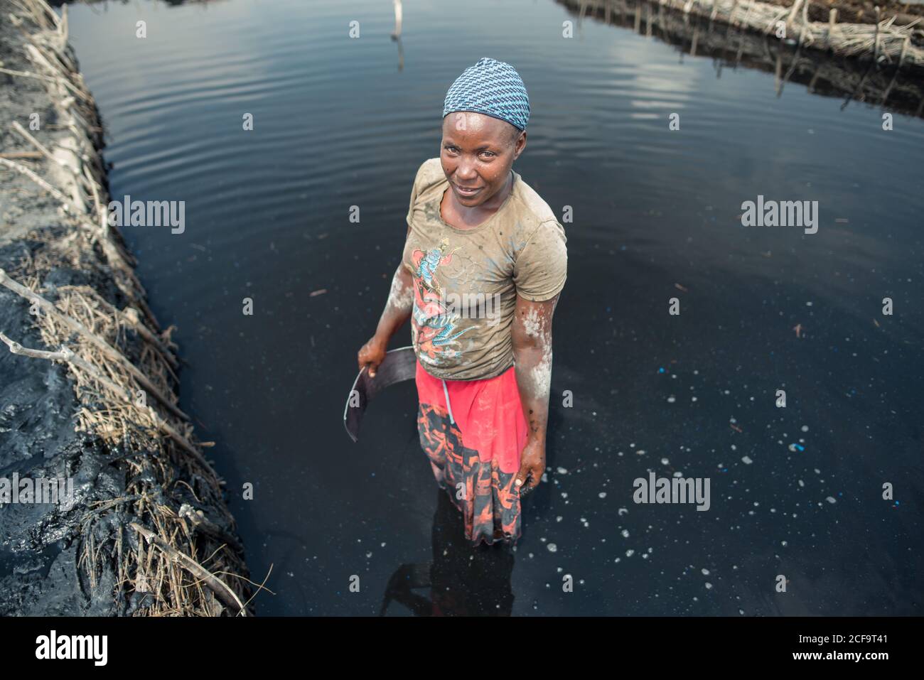 Uganda - 26 novembre 2016: Pensive sorridente donna africana in vividi abiti casual che mette a fuoco e minerando fango nero salato mentre si trova in piedi in acqua scura e utilizzando uno strumento metallico su saline farm contro la vista offuscata di piantagione e cielo blu chiaro in giornata calda Foto Stock