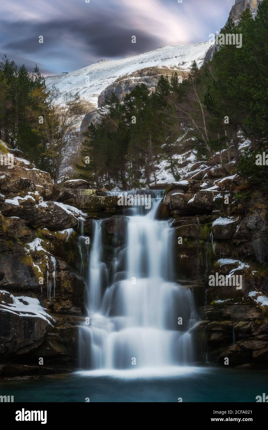 Vista panoramica incredibile della piccola cascata di montagna fredda che scorre attraverso scogliere innevate circondate da foreste di conifere in inverno Foto Stock