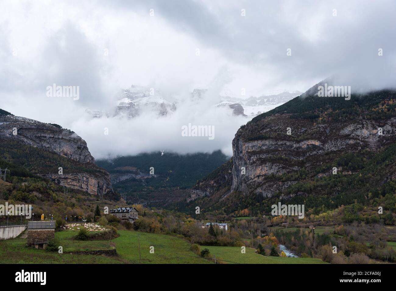 Maestose montagne rocciose e valle verde con piccole case di villaggio coperto di nebbia fitta e nuvole nel giorno d'autunno Foto Stock