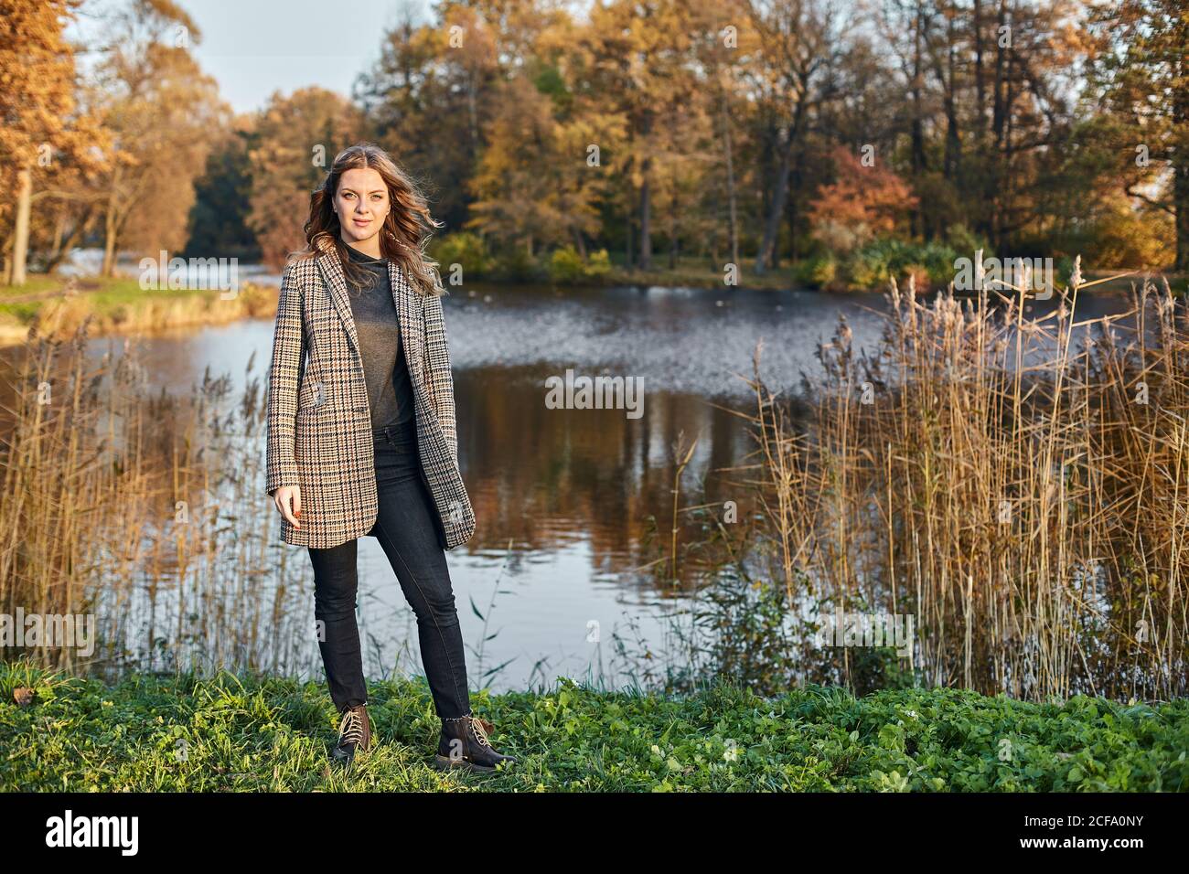 Donna sorridente dai capelli lunghi con cappotto marrone e in piedi in autunno parco su un lago Foto Stock