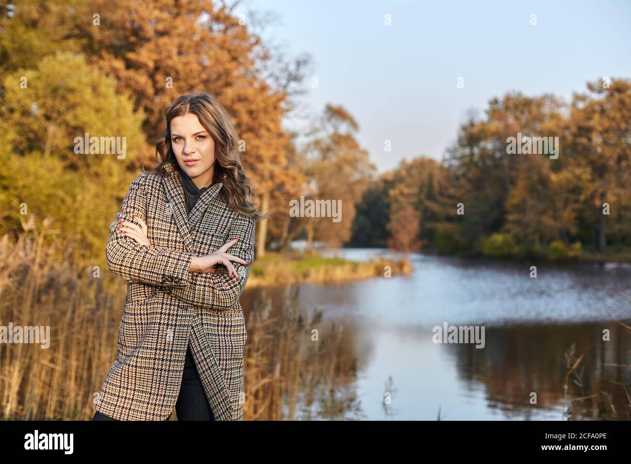 Donna dai capelli lunghi e serii che indossa un cappotto marrone e si alza in piedi in autunno parco su un lago Foto Stock