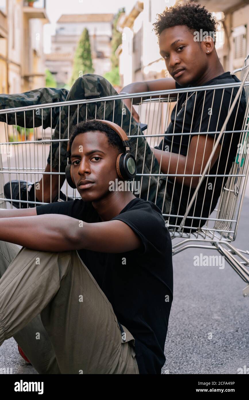 Felice giovane afroamericano uomo in vestiti casual seduto dentro cart con un amico maschio nero in cuffie durante il passatempo insieme in strada guardando la macchina fotografica Foto Stock