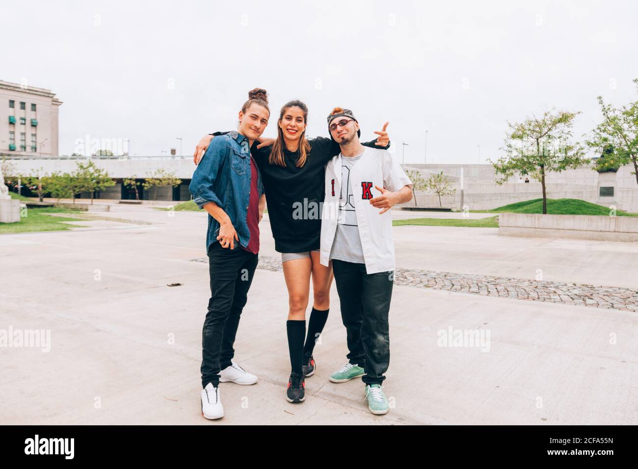 Amici di sesso maschile e femminile in un abbigliamento casual elegante in piedi con le braccia si aggirano mentre sorridi e guarda la fotocamera a vuota strada della città Foto Stock