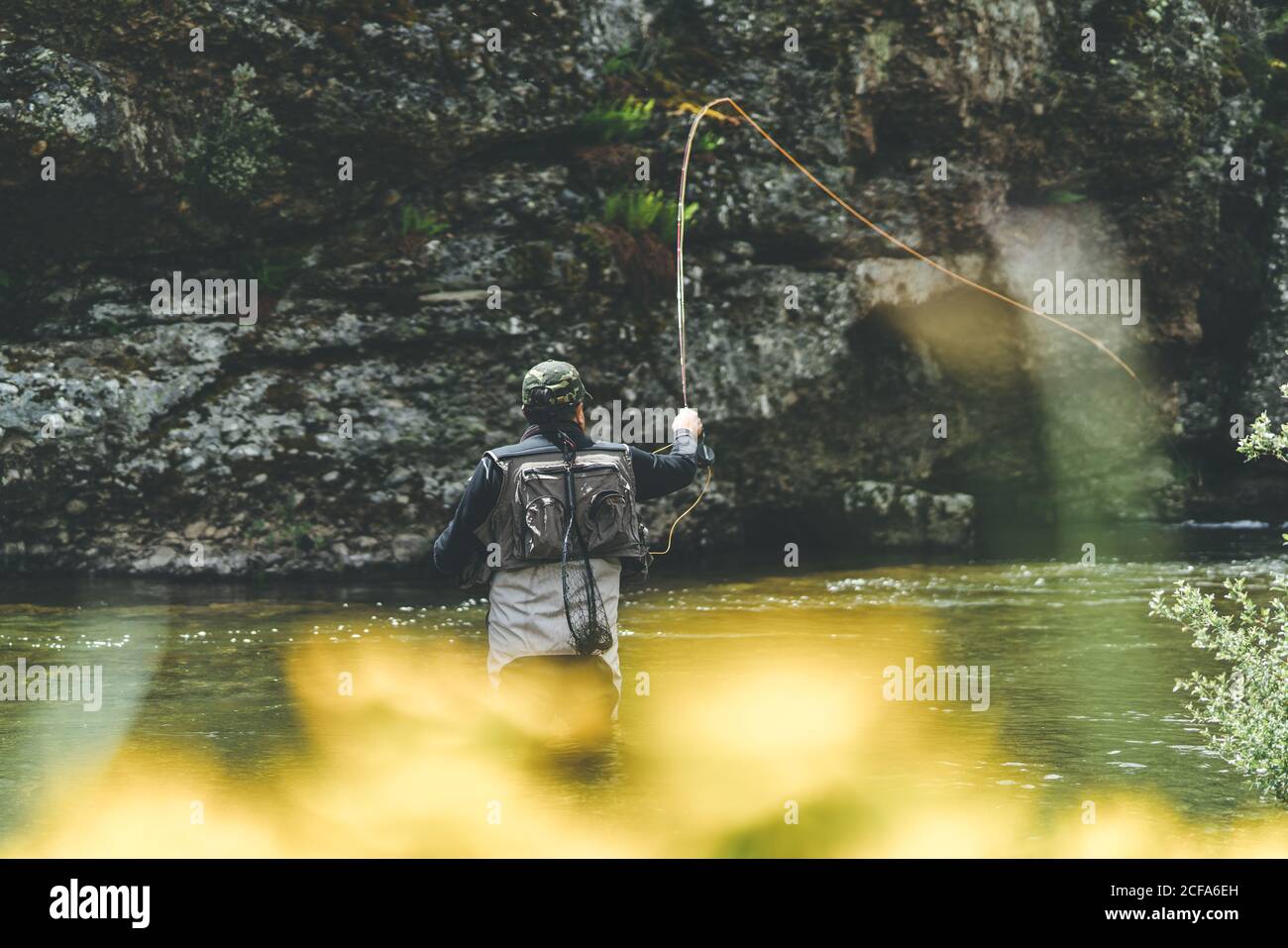 Vista posteriore del pesce harling uomo attrezzato mentre si è in piedi waders in torrente di montagna da scogliera e foresta Foto Stock