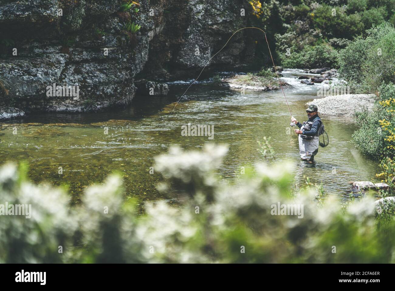 Vista laterale del pesce harling uomo attrezzato mentre si è in piedi waders in torrente di montagna da scogliera e foresta Foto Stock