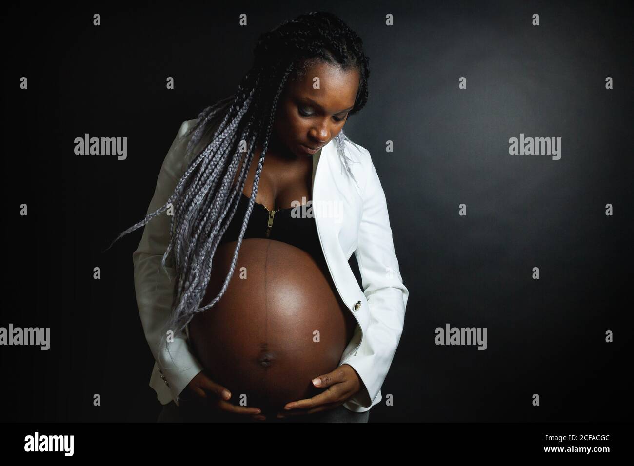 Giovane donna afro-americana incinta con trecce con blazer bianco e i jeans stroking pancia mentre si siede in studio contro nero parete Foto Stock