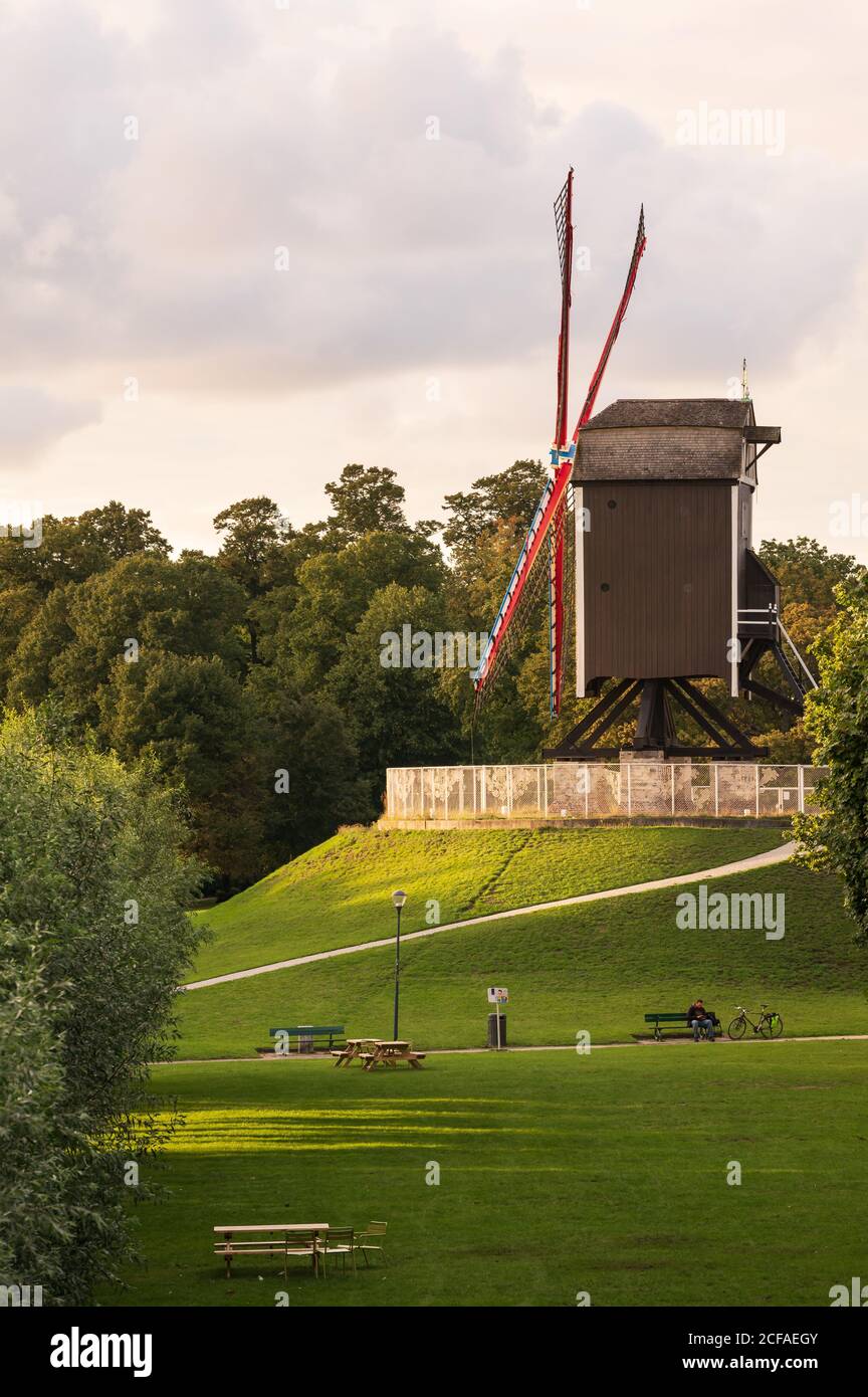 Immerso nella luce del tramonto, il mulino a vento Sint-Janshuis sorge su una piccola collina verde in un parco con panchine, lampade e alberi. Foto Stock