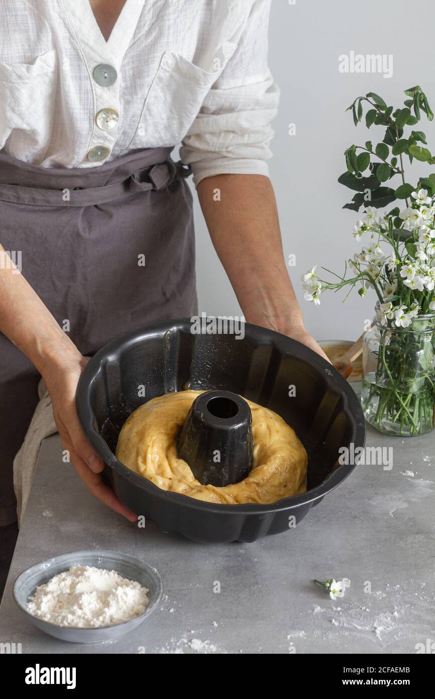 donna irriconoscibile in grembiule che mostra fuori rotolo di pasta morbida In una padella vicino alla farina e ai fiori mentre si prepara la torta Bundt sul tavolo Foto Stock
