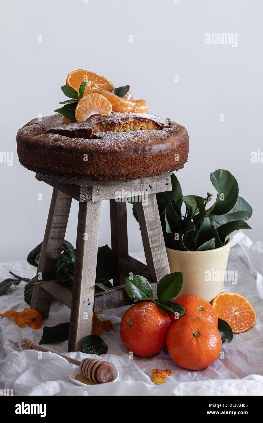 Pittoresca vita morta di fresco appetitoso taglio e tangerina intera torta fresca e gustosa su sgabello di legno e pianta verde in pentola Foto Stock