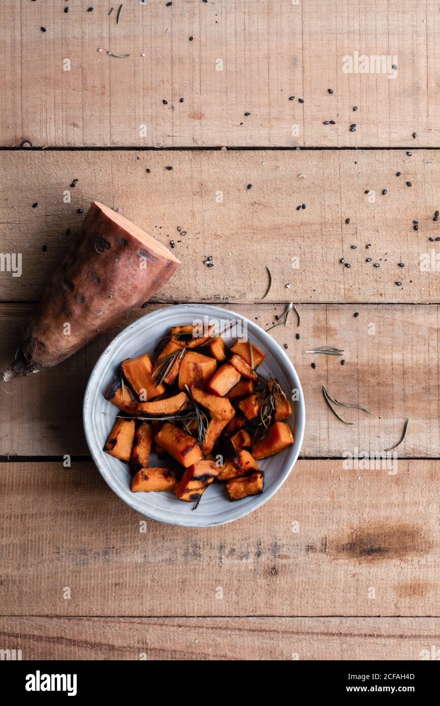 Vista dall'alto della metà delle patate dolci crude e dei pezzi di patate dolci al forno in ciotola su rustico tavolo di legno Foto Stock