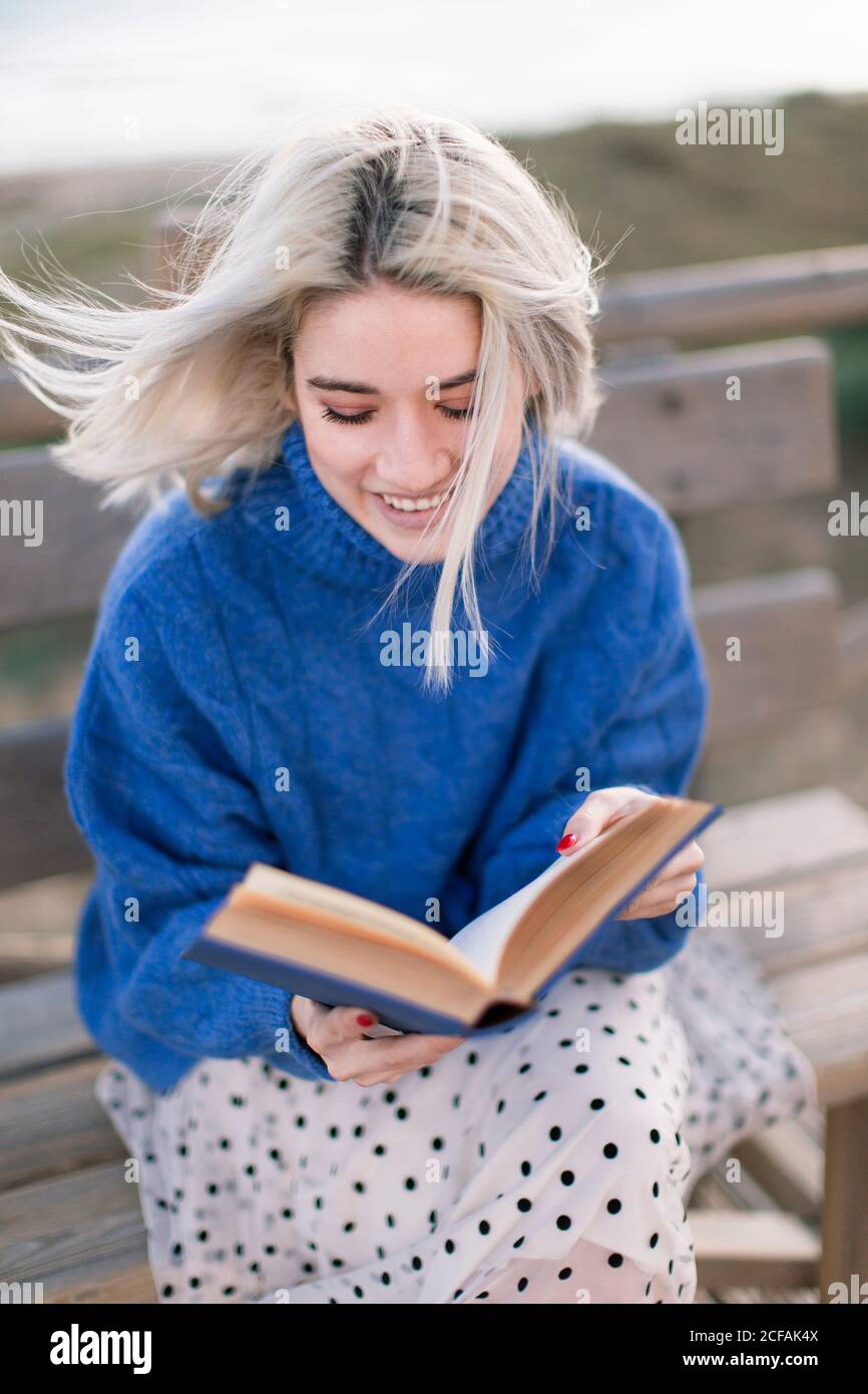 Giovane donna bionda con un caldo pullover blu e una gonna lontano mentre si siede su panca di legno in terrazza contro sfocato spiaggia e libro di lettura Foto Stock